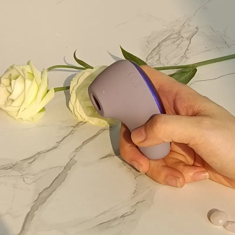 Stimulateur Virgule violet tenu en main, fond marbré avec rose